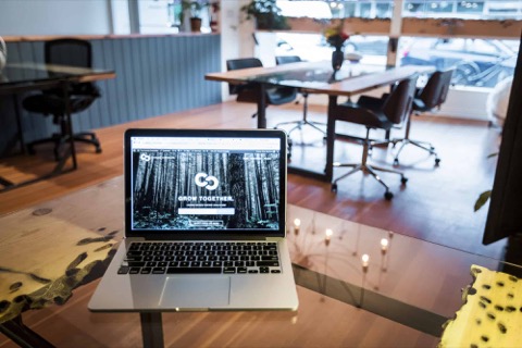 A laptop open on a table at The Common coworking space, with meeting tables, office chairs and tall windows behind