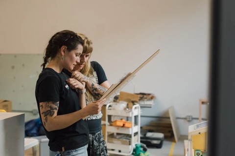 Two people at Create Makerspace looking closely at a laser-cut wooden panel, with shelves of materials behind them