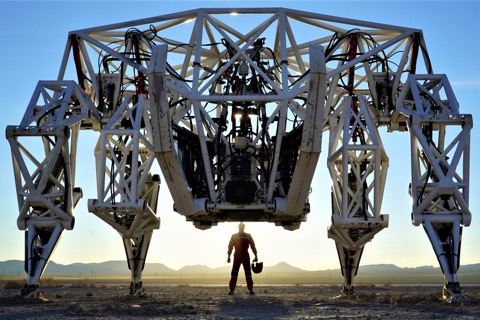Prosthesis, a huge white four-legged mech, standing in desert scrub at sunset with its pilot in a red suit beneath it for scale